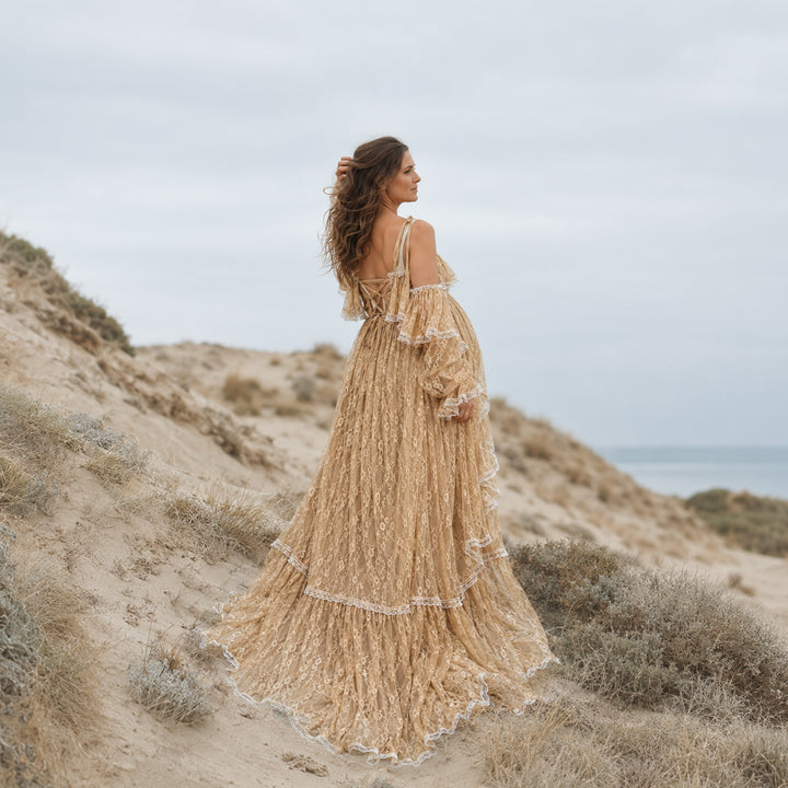 A pregnant woman stands on a sandy coastal dune wearing a Melinoe khaki  lace maternity gown with tiered ruffles, bell sleeves, and an open back with crisscross straps.