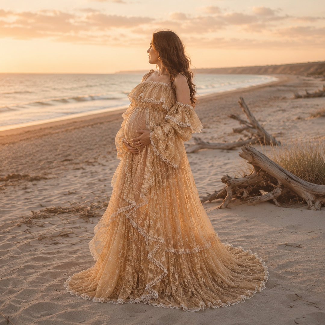 A pregnant woman stands on a sandy beach at sunset, wearing a Melinoe flowing lace maternity gown with off the shoulder ruffled sleeves and a long, tiered skirt that spreads across the ground