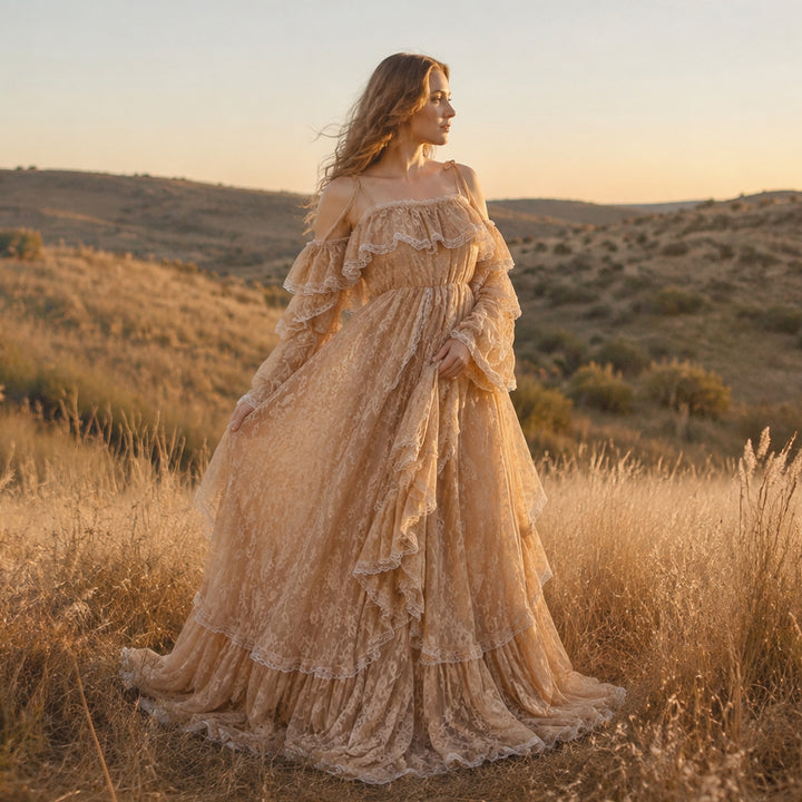A pregnant woman stands in a sunlit, dry grassy landscape wearing a Melinoe flowing lace maternity gown with off-the-shoulder ruffled sleeves and a tiered full skirt that spreads across the ground.