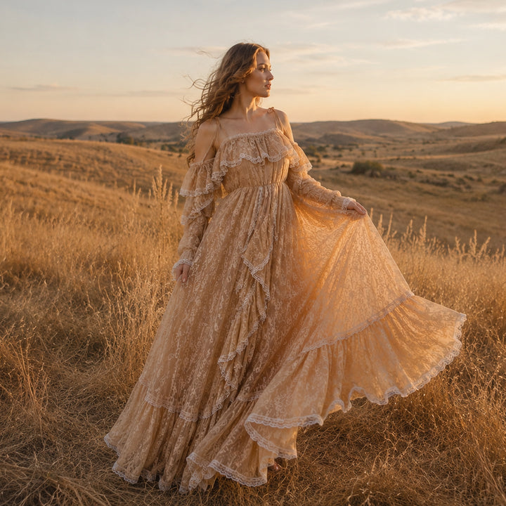 A pregnant woman stands in a sunlit, dry grassy landscape wearing a Melinoe flowing lace maternity gown with off-the-shoulder ruffled sleeves and a tiered full skirt that spreads across the ground.