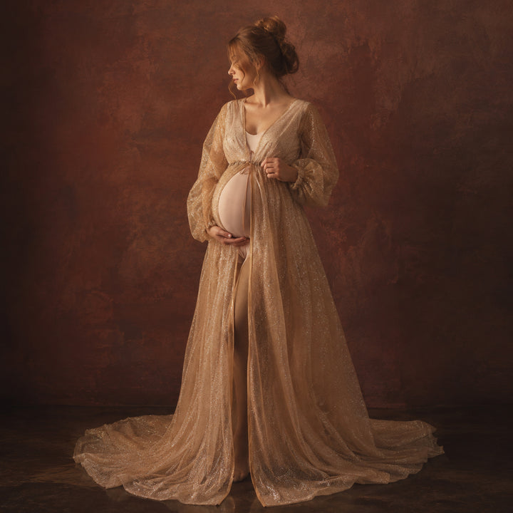 A pregnant model in a Metis khaki sequined V-neck robe stands against a dark red background, wearing a light bodysuit underneath with one hand on her belly; the robe features puff sleeves, a tie belt, and a floor-length train.