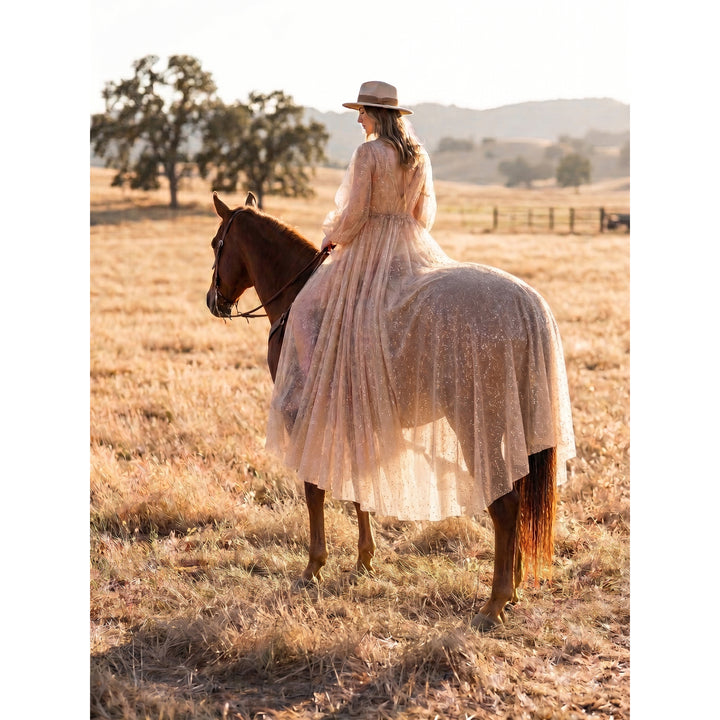 A hat-wearing woman in a Metis sequined gown rides a brown horse in dry grass.
