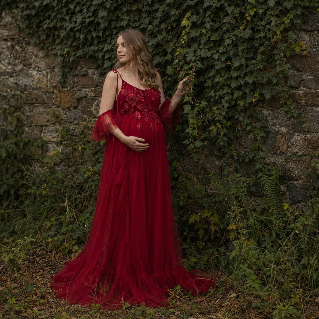 A pregnant woman wears a Oreades red dress with 3D floral embroidery and ruffled sleeves, it's train trailing on the ground; she touches ivy with her left hand, cradles her belly with her right, standing.
