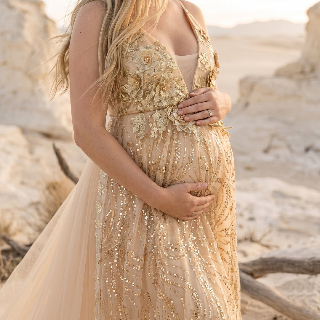 A pregnant model in a Pandia gold sequined spaghetti strap gown holds her belly; the dress features 3D floral embroidery and sequins, with desert rocks in the background.