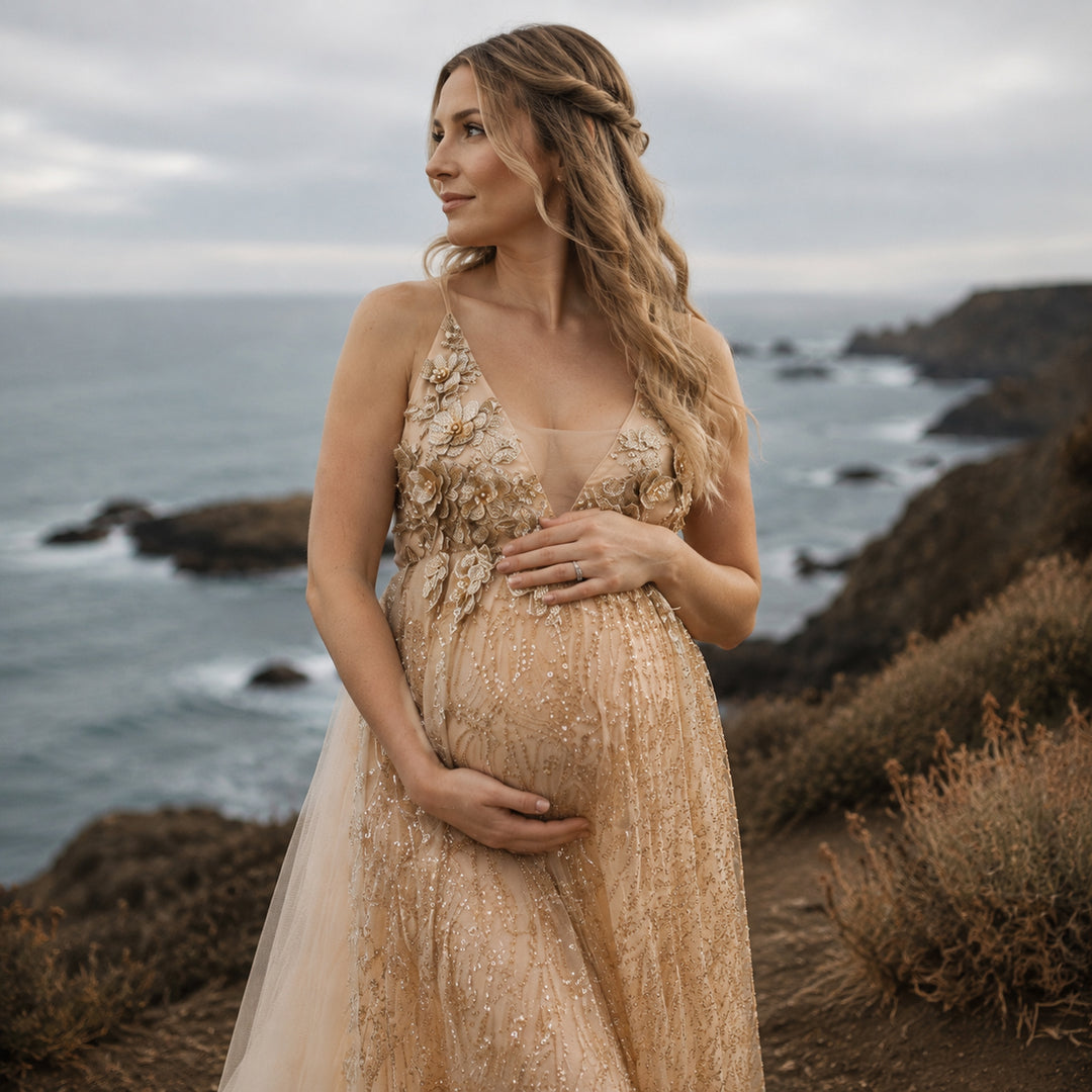 A pregnant model in a Pandia gold sequined spaghetti strap gown stands on a seaside cliff, hands on belly.