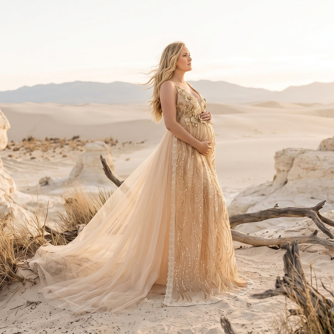 A pregnant model in a Pandia gold sequined spaghetti strap gown stands in the desert, hands on belly.