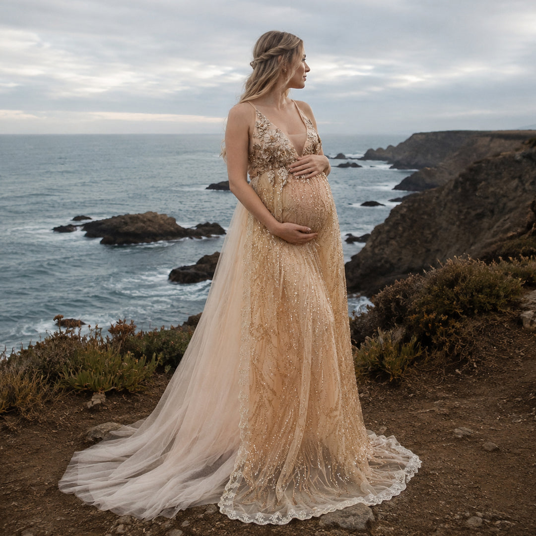 A pregnant model in a Pandia gold sequined spaghetti strap gown stands on a seaside cliff, hands on belly.