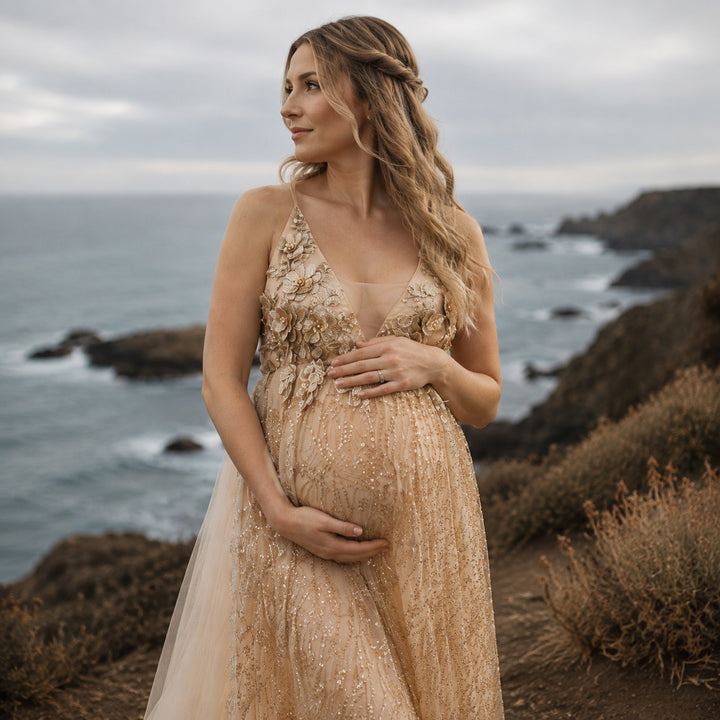 A pregnant model in a Pandia gold sequined spaghetti strap gown stands on a seaside cliff, hands on belly.