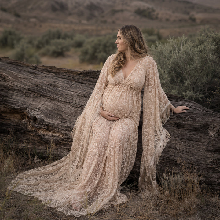 A pregnant woman in a Pheme beige lace fringe gown sits sideways on wilderness deadwood, hands on belly.