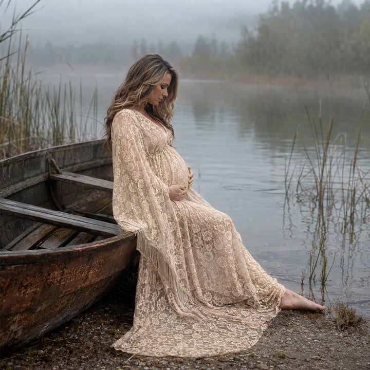 A pregnant woman in a Pheme  khaki lace fringe gown sits by an old wooden boat at a lakeside, hands on belly.