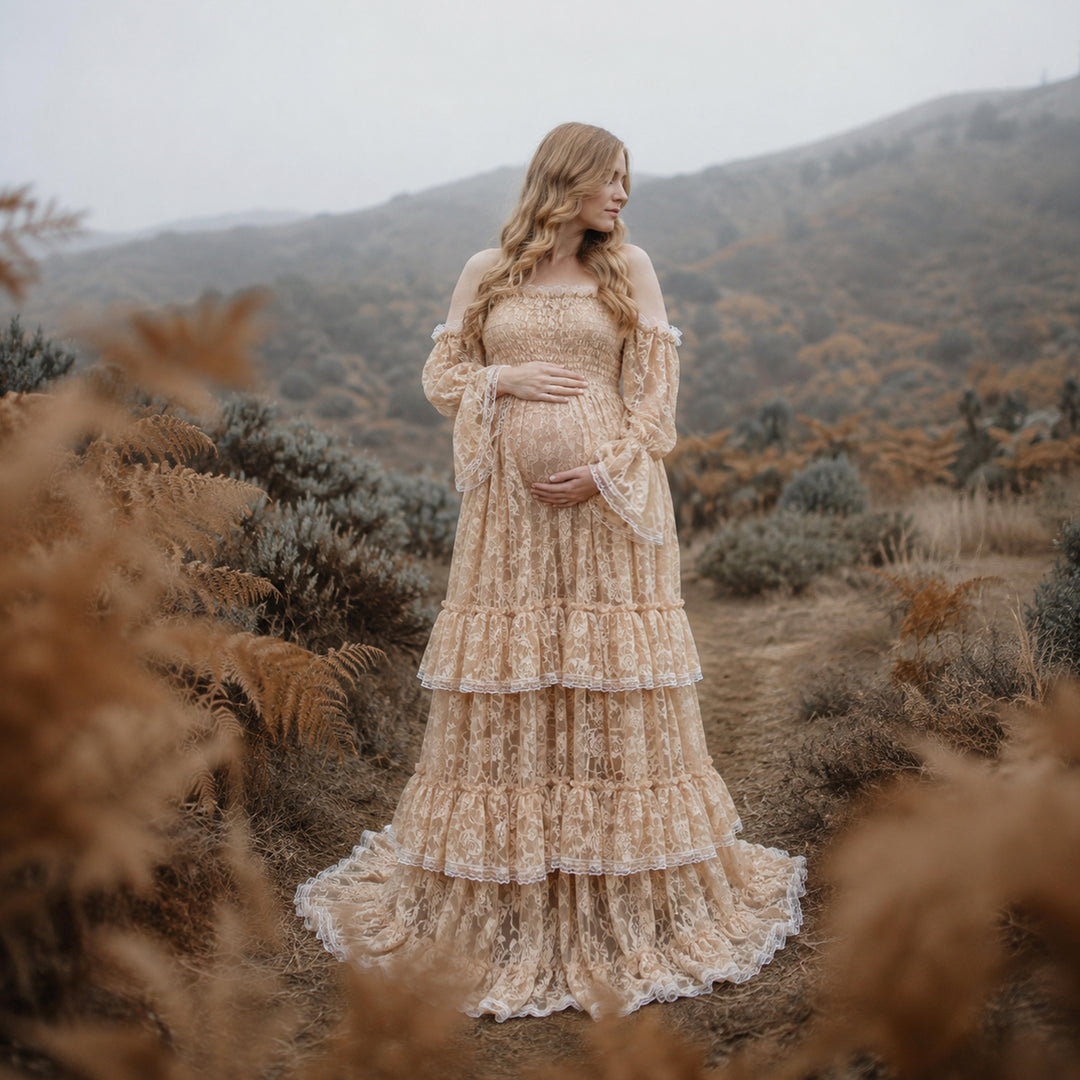 A pregnant woman in a Selene khaki lace off-shoulder gown stands in the wild; the dress features bell sleeves, a tiered ruffled hem, and a train. She cradles her belly with both hands.