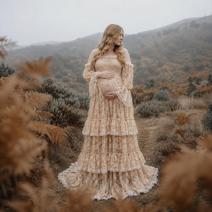 A pregnant woman in a Selene khaki lace off-shoulder gown stands in the wild; the dress features bell sleeves, a tiered ruffled hem, and a train. She cradles her belly with both hands.