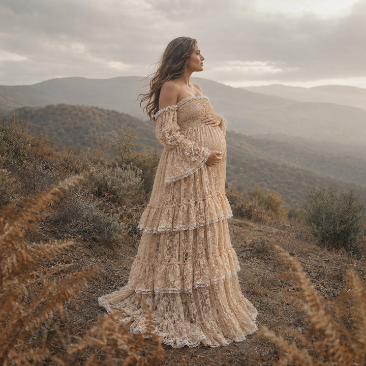 A pregnant woman in a Selene khaki lace off-shoulder gown stands in the wild; the dress features bell sleeves, a tiered ruffled hem, and a train. She cradles her belly sideways with both hands.
