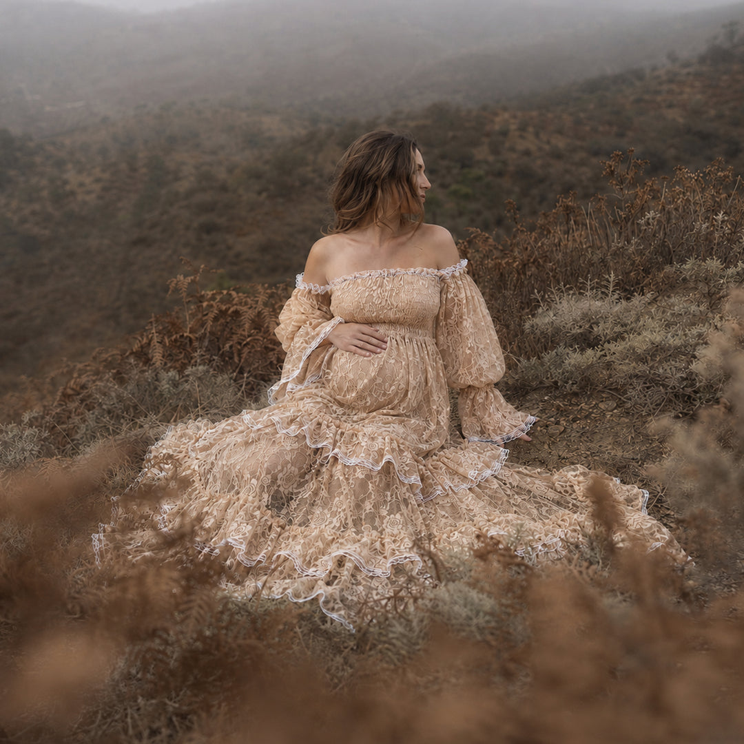 A pregnant woman in a Selene khaki lace off-shoulder gown sits in the wild; the dress features lantern sleeves, a tiered ruffled hem, and a train. She cradles her belly sideways with one hand.