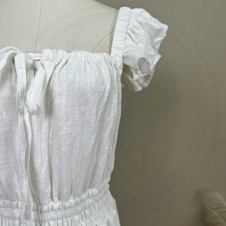 A ivory Tethys gown is displayed on a mannequin, featuring ruffled short sleeves, a front tie, and an elasticated waist.