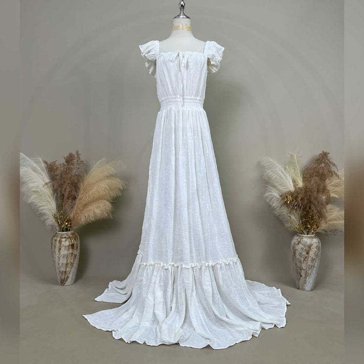A ivory Tethys gown is displayed on a mannequin with ruffled short sleeves, a front tie, and an elasticated waist; it features a tiered ruffled hem with a train. Ceramic vases with dried flowers are placed on both sides.