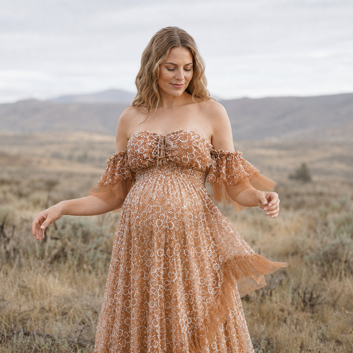A pregnant woman in a Tyche khaki sheer off-shoulder gown with white embroidery; she looks down with hands hanging naturally.