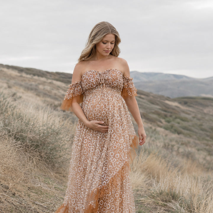 A  pregnant woman is wearing Tyche khaki stands on hills and dry vegetation; she looks down, cradling her belly with her right hand.