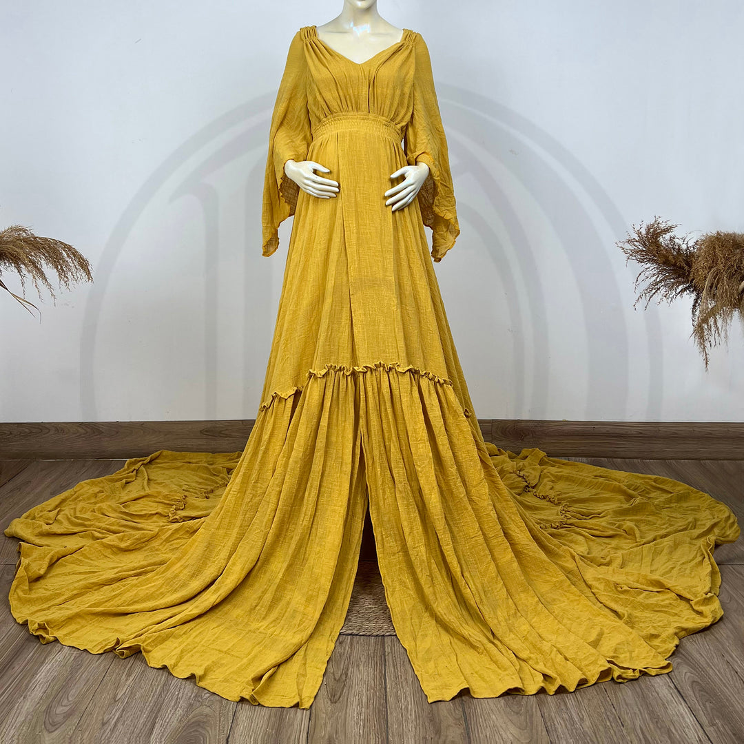 A Daphne yellow long-sleeved V-neck gown is displayed on a mannequin, with hands positioned on the simulated belly. The dress features a high front slit and its hem and train are spread out on the wooden floor.