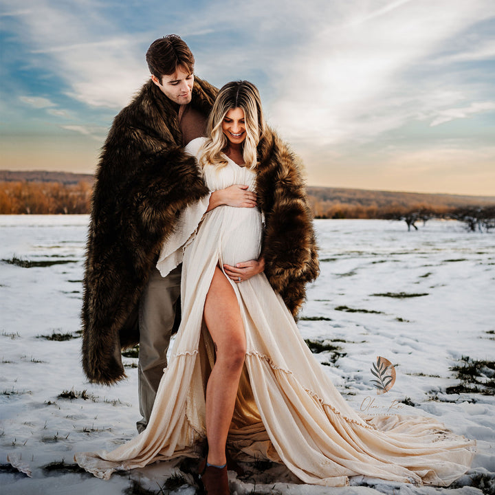 A man in a fur coat stands behind a pregnant woman in a Daphne slit dress in the snow, hands on her belly. A watermark is on the right.