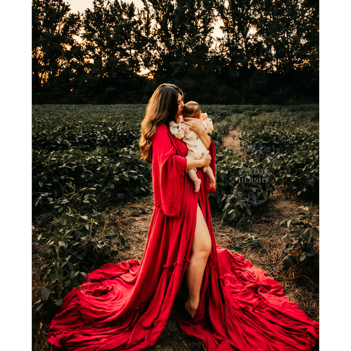 a woman in a Daphne red slit dress stands barefoot among plants, holding an infant. A text watermark appears on the right.