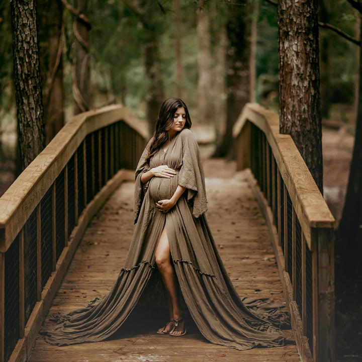 A pregnant woman in a Daphne olive long dress with a slit stands on a wooden bridge in a forest, holding her belly.